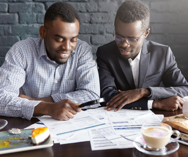 Two handsome successful African-American businessmen using magnifying glass while studying documents and reviewing finances, sitting at cafe table with papers, having happy cheerful expressions